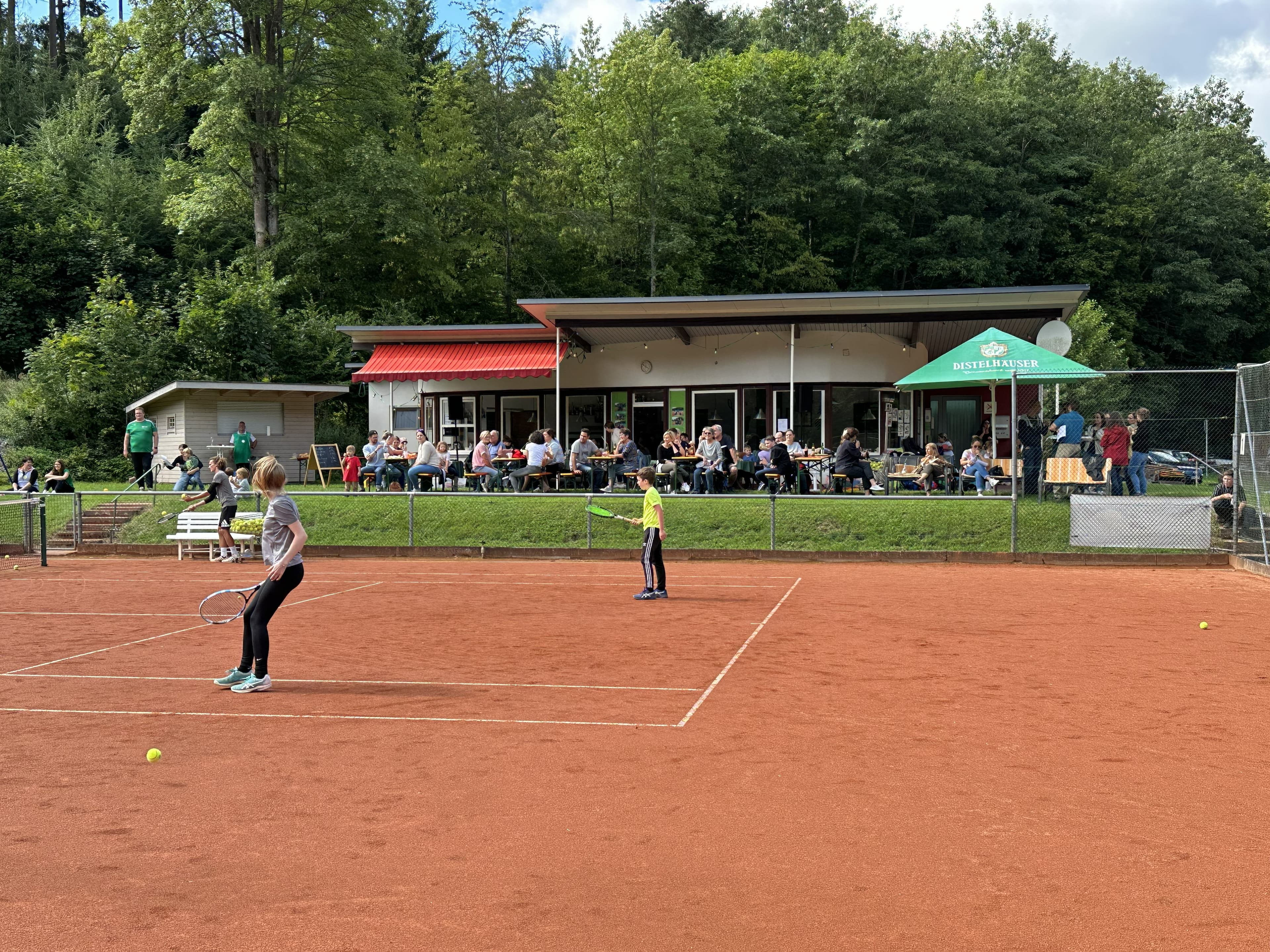 Clubhaus des TC Grün-Weiß Buchen bei einem Vereinsnachmittag — voll besetzte Terrasse und Kinder beim Training auf dem Sandplatz